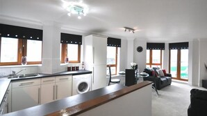 Egy modern konyha mosógéppel, egy étkező asztallal és székekkel, valamint nagy ablakokkal, melyekre fekete árnyékolók vannak felszerelve.