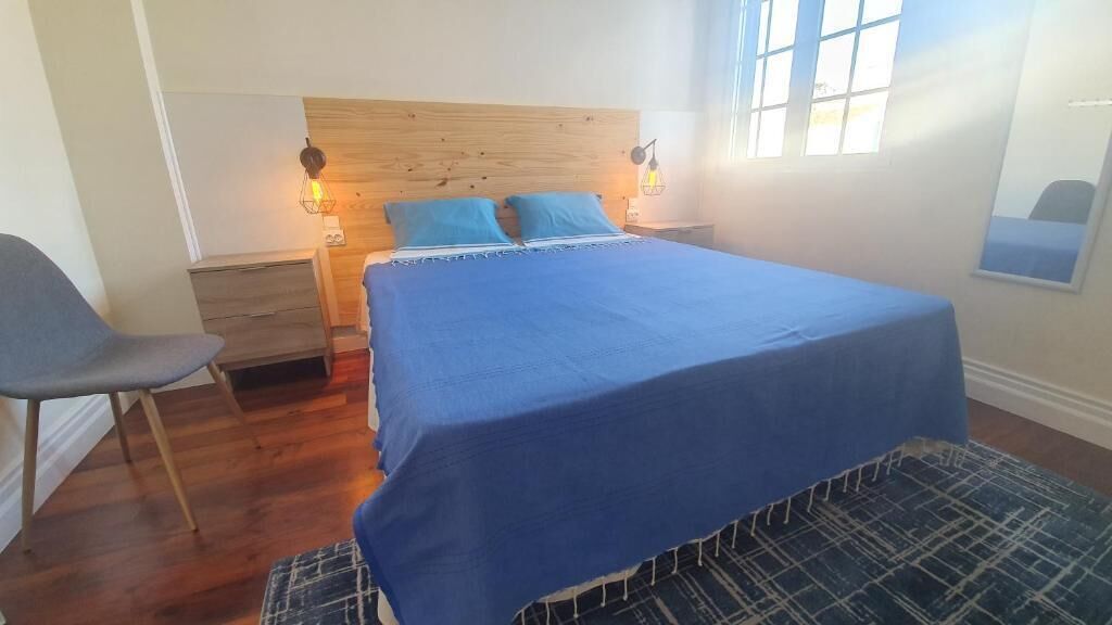 A bedroom with a wooden headboard, a blue bedspread, two pillows, a chair, a nightstand, and a lamp.