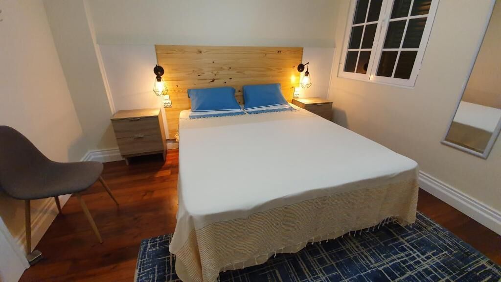 A bedroom with a wooden headboard, a bed with blue pillows, bedside tables with lamps, a chair, and a window.