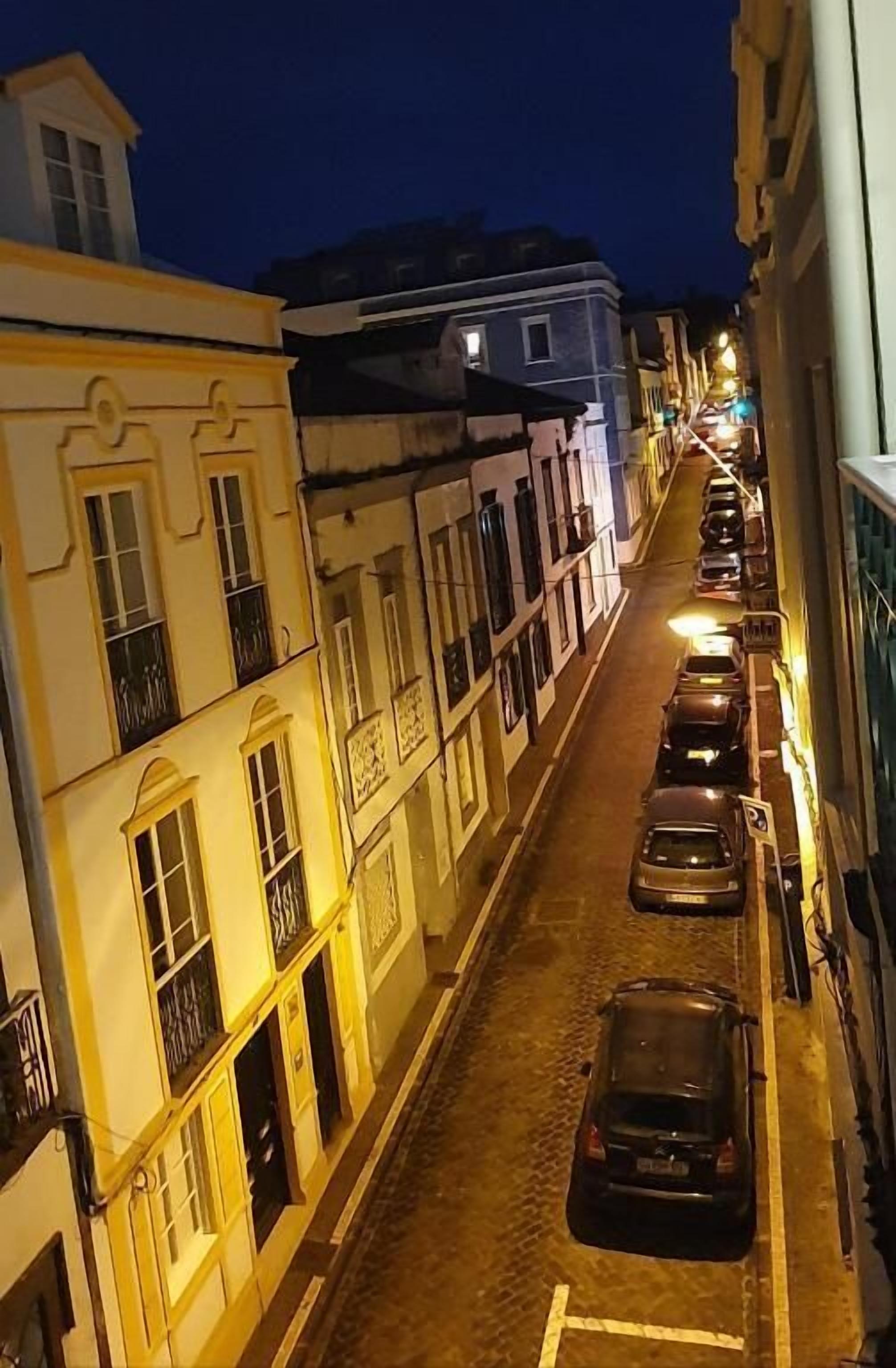 A narrow cobblestone street lined with historic buildings, parked cars, and a clear sky.