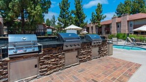 An outdoor barbecue area with multiple grills, a stone wall, and a poolside lounge with umbrellas. - Cozy Casa | Executive Condo•Pool•Spa•Sauna•BBQ (San Diego)