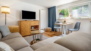 Una sala de estar moderna con un televisor de pantalla plana, un mueble de madera, una mesa de centro de vidrio, dos sillones y un ventanal con vistas a la vegetación.