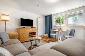 Ein modernes Wohnzimmer mit einem Flachbildfernseher, einer Holzkommode, einem Glastisch, zwei Stühlen und einem großen Fenster mit Blick auf Grünflächen.