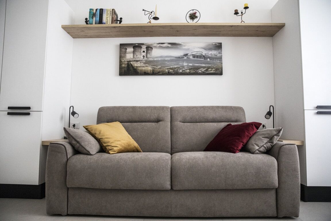 A grey sofa with two cushions, a wooden shelf above with books and decorative items, and a framed picture of a castle on the wall.