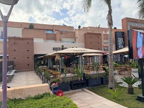 Zona de café al aire libre con mesas y sombrillas, rodeada de follaje y un edificio de diseño moderno.