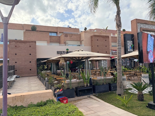 Un espace café en plein air, avec des tables et des parasols, entouré de verdure et bordé d’un bâtiment au design moderne.
