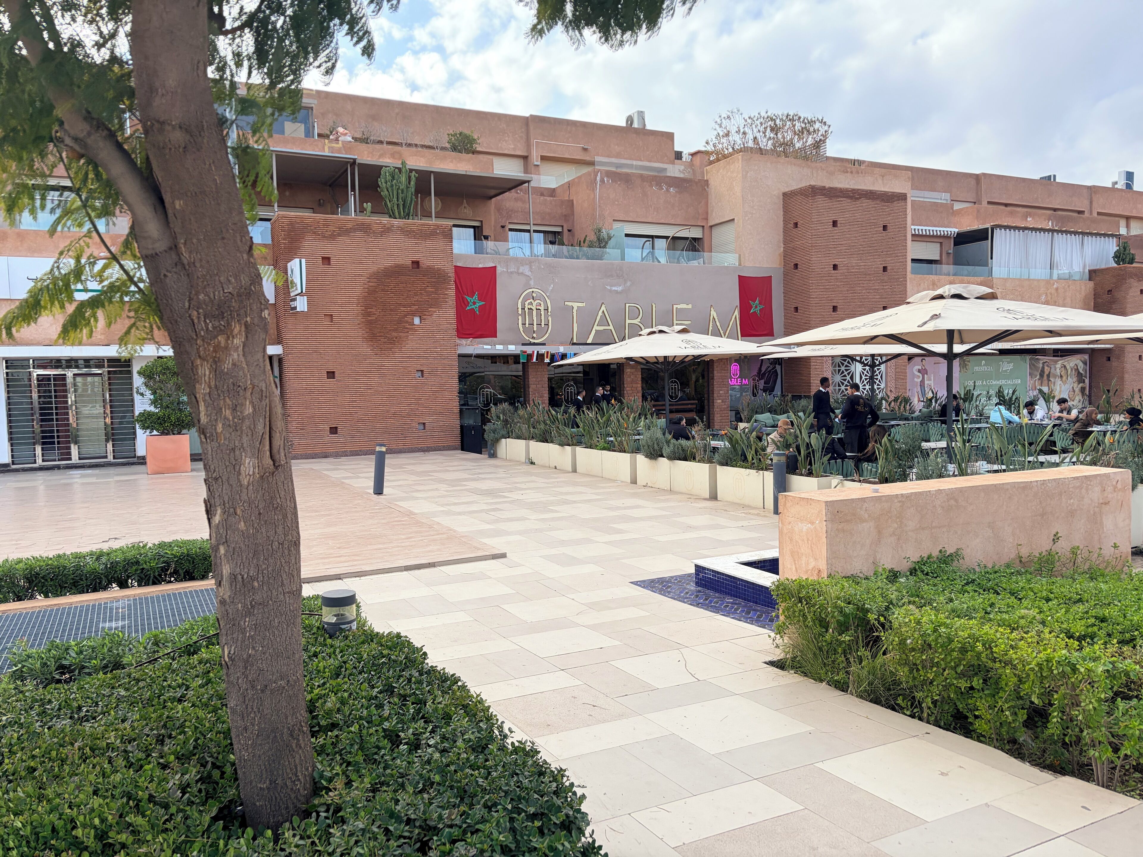 A courtyard with outdoor seating, a restaurant named "Table M," and a building with a Moroccan flag.