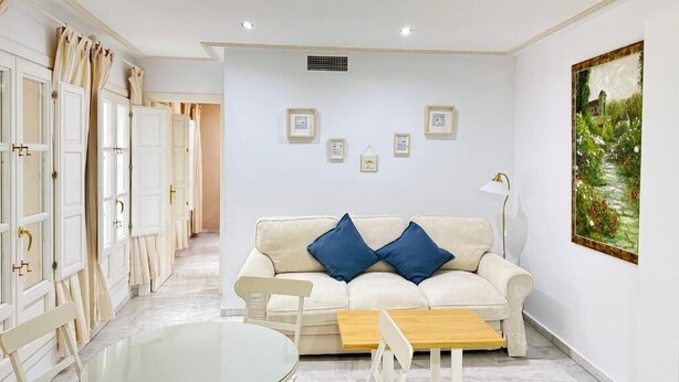 Un salon moderne avec un canapé blanc, deux coussins bleus, une table basse en verre et un tableau accroché au mur.