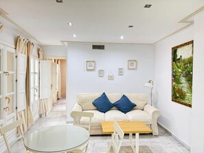 Uma sala de estar moderna com um sofá branco, duas almofadas azuis, uma mesa de centro de vidro e uma pintura na parede.