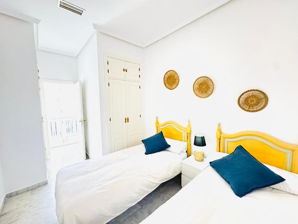 Une chambre avec deux lits, du linge de lit blanc et une tête de lit jaune. On y trouve trois décorations murales tissées et une porte blanche avec une poignée.