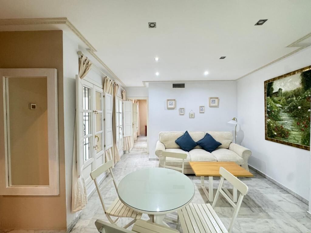 Une pièce avec un canapé blanc, une table ronde en verre et une table basse en bois. Des tableaux encadrés ornent le mur, et un tableau est accroché au-dessus du canapé.