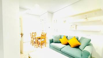 A living room with a teal sofa, a white coffee table, and yellow cushions. There is a dining area with wooden chairs and a table.