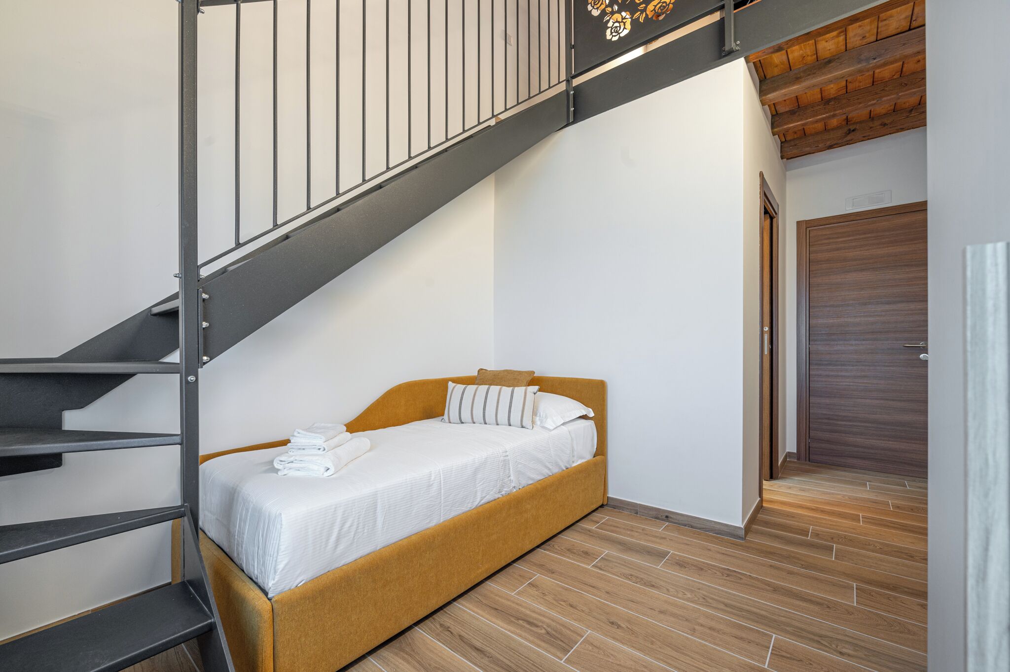 A single bed in a room with a wooden floor, a staircase with metal railings, and a wooden door.