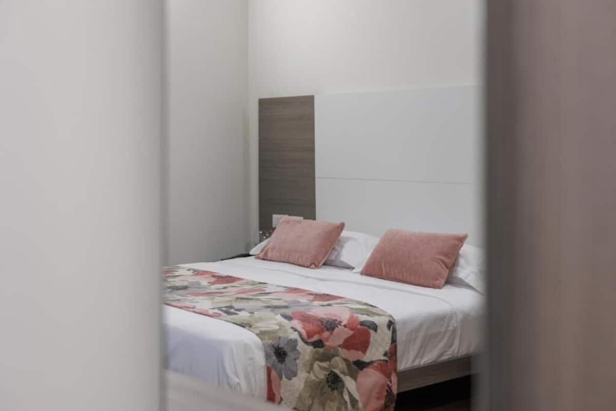 Un lit simple avec une couvre-lit à motifs floraux et deux oreillers roses, dans une pièce dont un mur est d’un blanc uni et qui possède une tête de lit en bois.