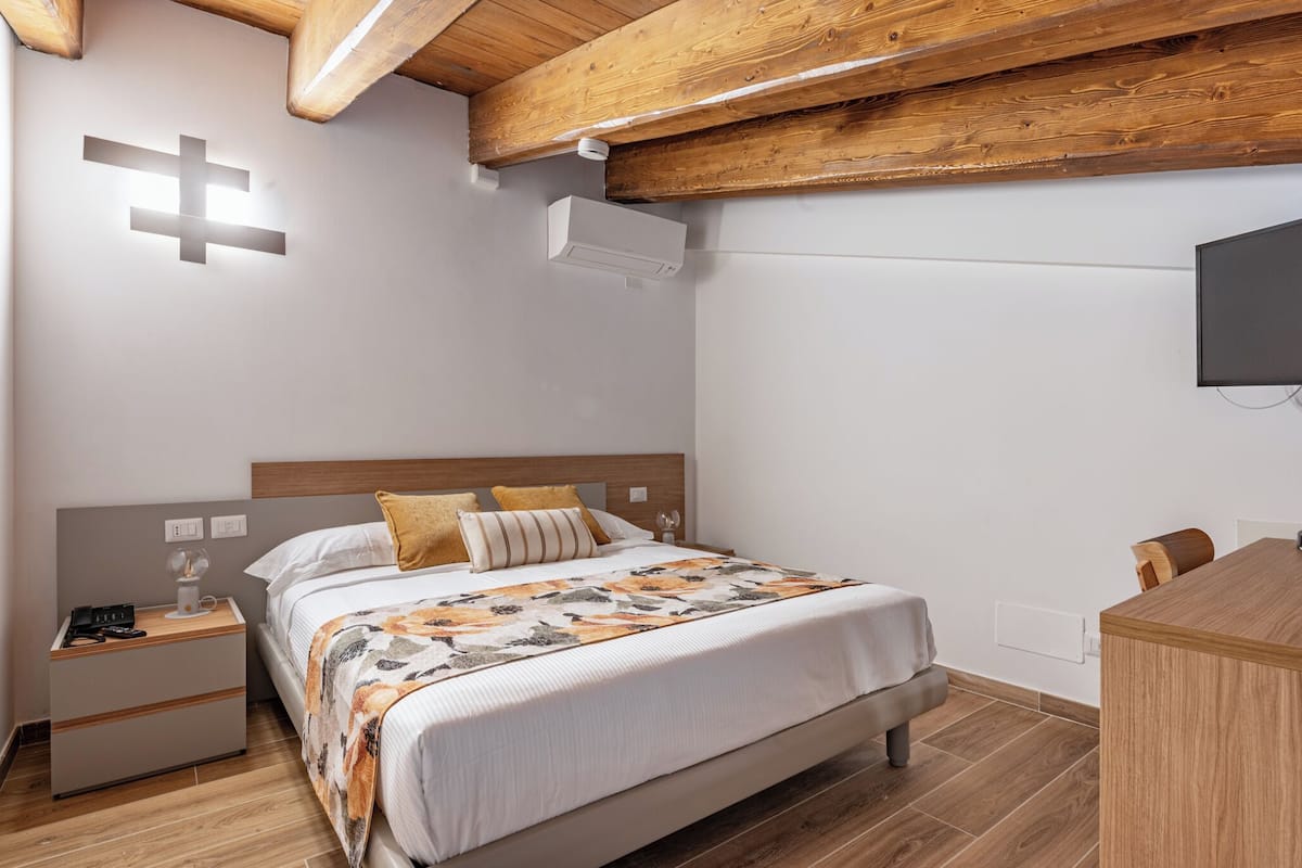 Une chambre avec un plafond en bois, un lit recouvert d’une couvre-lit à motifs, une table de chevet avec une lampe et un bureau avec une chaise.