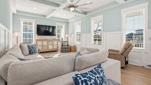 Un salon spacieux avec un grand canapé beige, un fauteuil beige et un ventilateur de plafond. On y trouve plusieurs fenêtres avec des volets blancs, un téléviseur à écran plat fixé au mur et un meuble en bois orné d’objets décoratifs.
