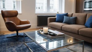 Ein modernes Wohnzimmer mit Ledersessel, Glastisch mit Kerze, Buch und blaues Sofa mit Kissen.
