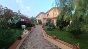 A paved pathway leading to a building with a beige facade, surrounded by lush greenery and flowering plants. - Tenuta Albachiara (Naso)