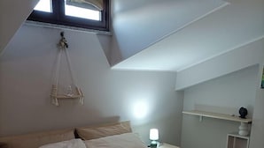 A bedroom with a bed, pillows, a bedside table with a lamp, a hanging macrame swing, and a window with curtains. - Tenuta Albachiara (Naso)