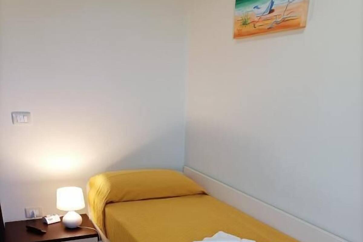 A small, single-bed room with a yellow bedspread, a bedside lamp, and a nightstand with a lamp.