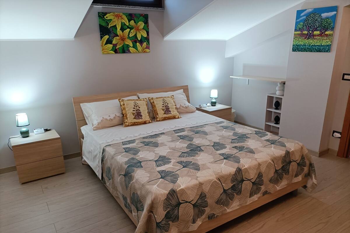 A bedroom with a bed, nightstands, a lamp, a painting on the wall, and a shelf with decorative items.