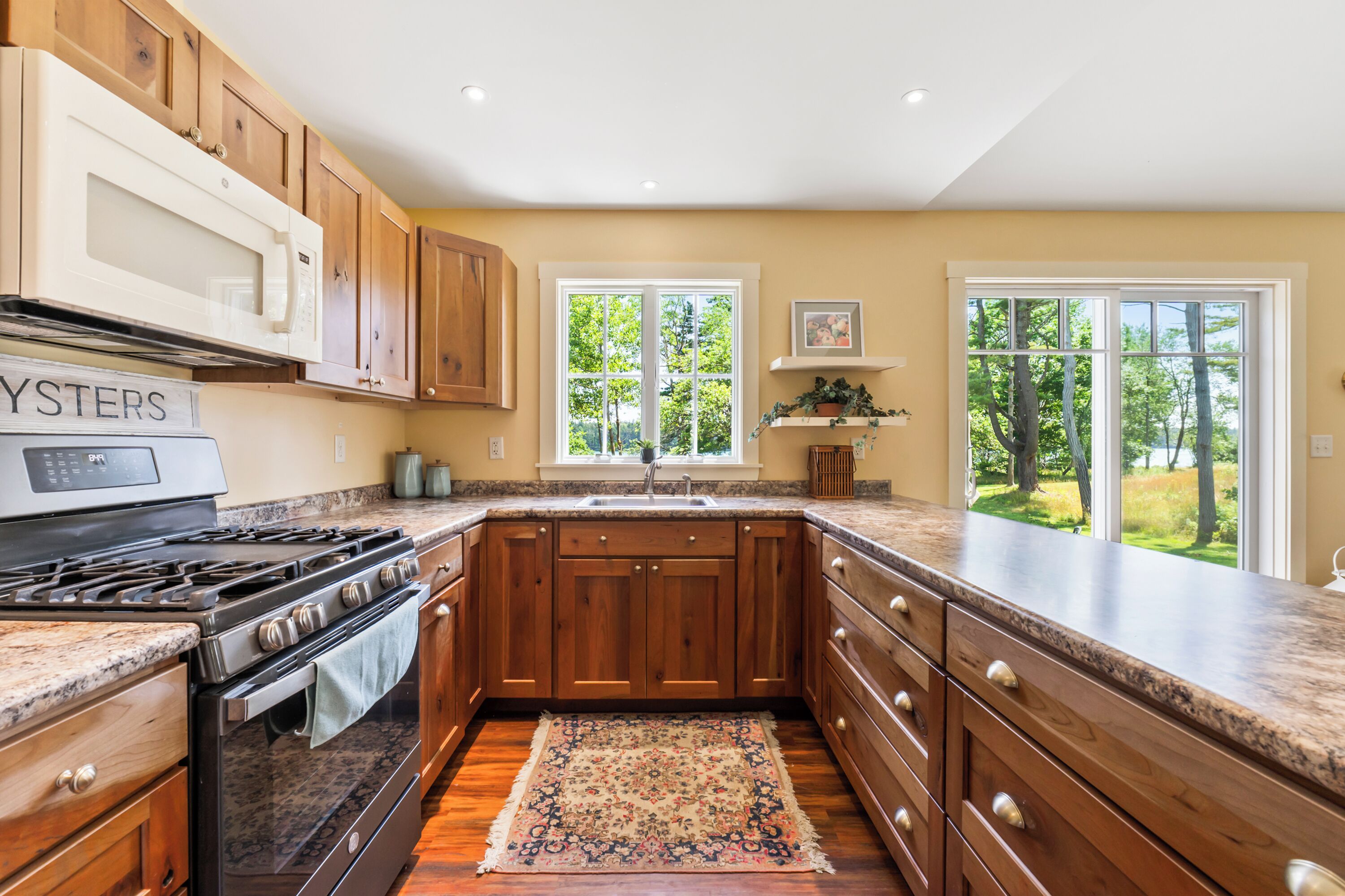 A kitchen with wooden cabinets, a stainless steel stove, granite countertops, and a large window with a view of trees.