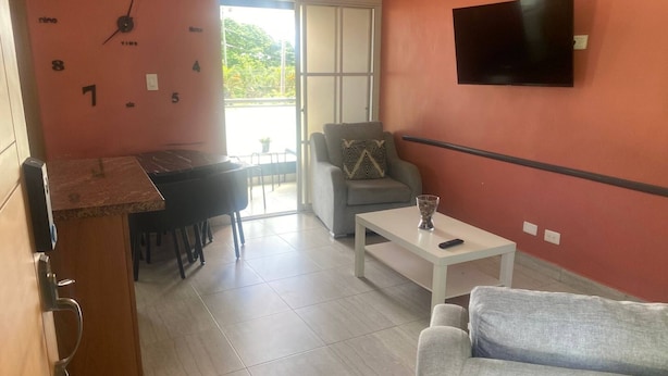 A living room with a grey sofa, a coffee table, a grey armchair, a TV mounted on the wall, and a kitchen area with a clock on the wall. - Modern 1 Bedroom Apt 5B (Puerto Plata)