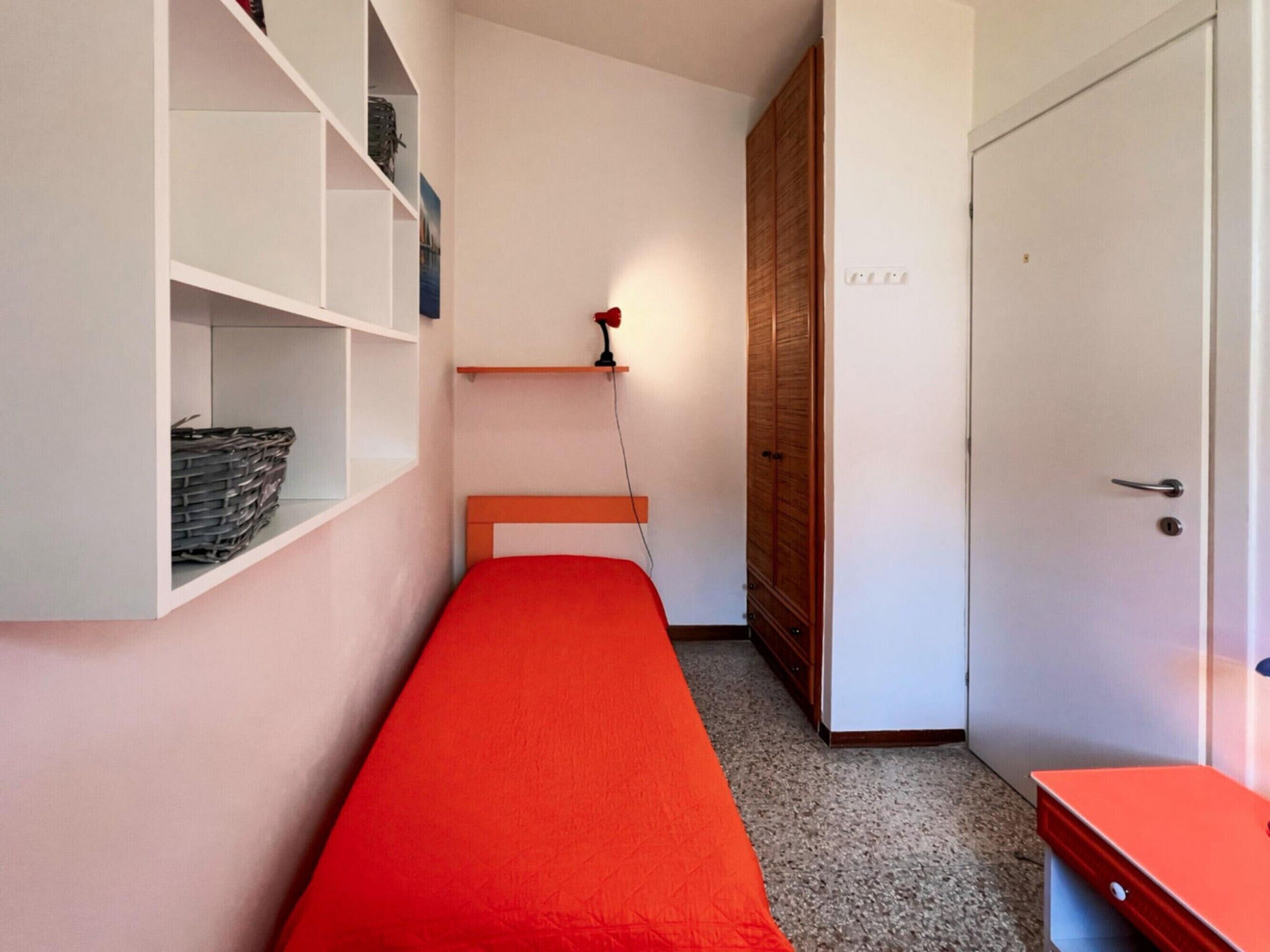 A small, narrow room with a single bed, a wardrobe, and a desk.