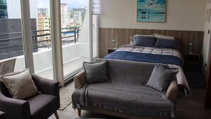 Un dormitorio con una cama grande, un sofá, un cuadro y vistas de la ciudad desde un balcón.