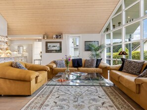 A spacious living room with a high ceiling, a glass coffee table, two leather sofas, and a large window offering a view of greenery.