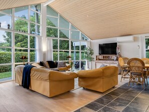 A spacious living room with a large glass wall, a sofa set, a dining area with wicker chairs, and a flat-screen TV mounted on the wall.