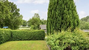Un jardín cuidado con un alto árbol de coníferas, setos bien recortados y un muro de piedra.