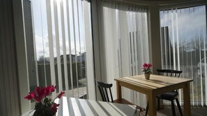 Een kamer met een tafel en stoelen, grote ramen met zonwering en uitzicht op buiten.