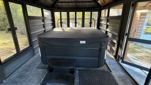 En utomhusbubbelpool med tak och en bänk, omgiven av stora fönster som ger utsikt över ett skogsområde.