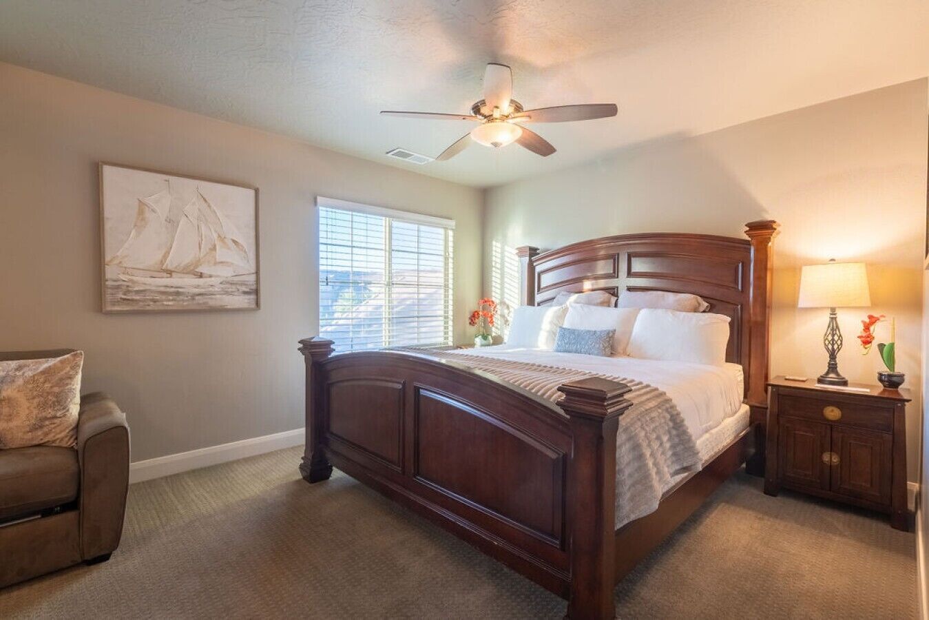 A bedroom with a wooden bed, a ceiling fan, a window with blinds, a nightstand with a lamp, and a chair.