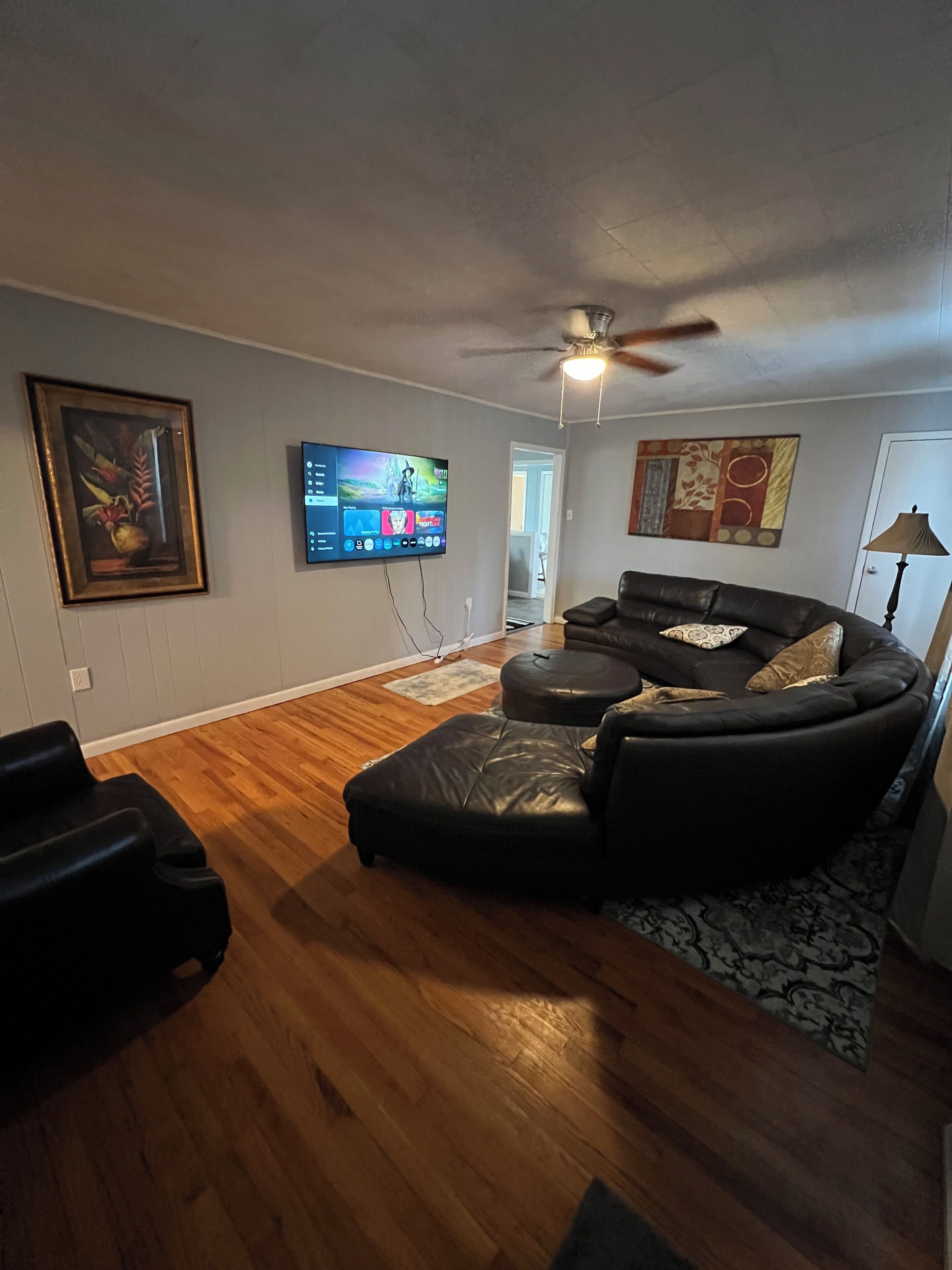 A living room with a black leather sofa, a flat-screen TV mounted on the wall, wooden flooring, and a ceiling fan.