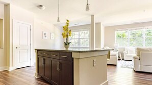 Uma sala de estar ampla com piso de madeira grande, uma ilha de cozinha marrom escura com um vaso de flores amarelas, e um sofá branco ao fundo.