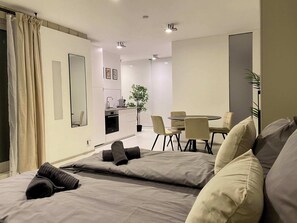Ett modernt hotellrum med en säng, en matplats med stolar, ett litet kök och en krukväxt.