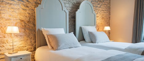 Una habitación con dos camas, cada una con cabecera, mesita de noche y lámpara, y una pared de piedra.