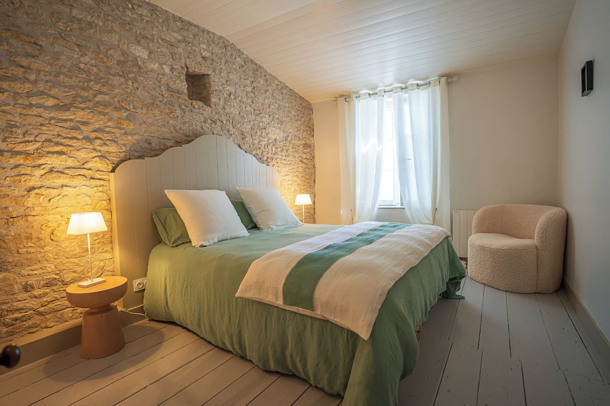 Un dormitorio con pared de piedra, cabecera de madera, colcha verde, lámpara de noche y una silla.