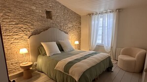 Un dormitorio con pared de piedra, suelo de madera, una cama con colcha a rayas verdes y blancas, dos lámparas de noche, una silla y una ventana con cortinas.