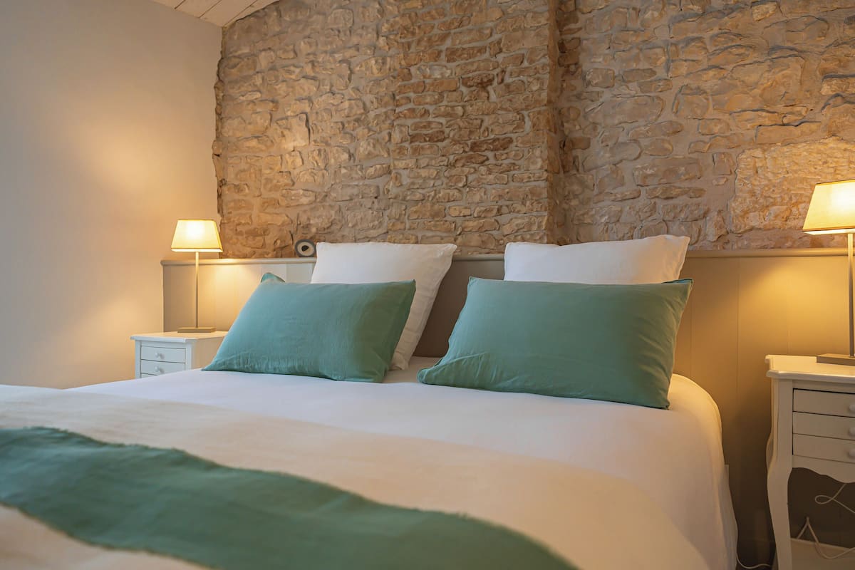 Una cama con dos almohadas, una mesita de noche con lámpara y una pared de piedra.