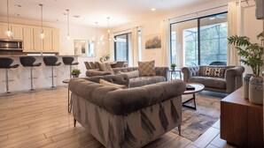 En moderne stue med sofa, lænestole og sofabord. Der er et køkkenområde med mikroovn og barstole.