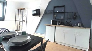 Ett kompakt kök med vita skåp, en mikrovågsugn och ett litet kylskåp. Det finns ett matbord med tallrikar och en kopp. En tv är monterad på väggen, och en hylla med köksartiklar syns.