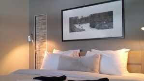 Una cama bien hecha con sábanas blancas y dos toallas negras, una fotografía en blanco y negro enmarcada en la pared, y una lámpara junto a la cama.