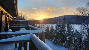Una cabaña de madera con una terraza cubierta de nieve y vistas a un paisaje nevado con un atardecer.