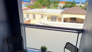 Ein Balkon mit zwei Stühlen, einem kleinen Tisch, einem Topfpflanzen und Blick auf Gebäude und einen klaren Himmel.