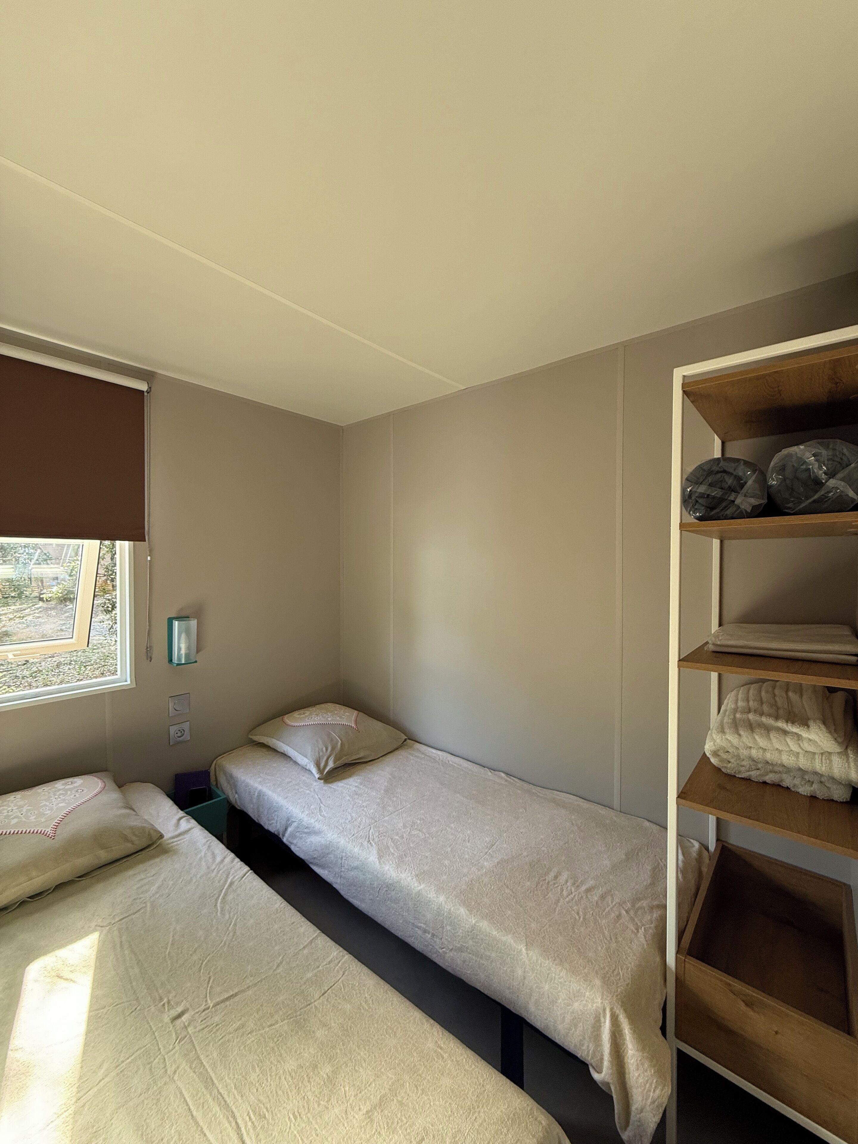 Una habitación pequeña y bien iluminada con dos camas, una ventana con persiana, un estante con toallas dobladas y una lámpara fijada a la pared.