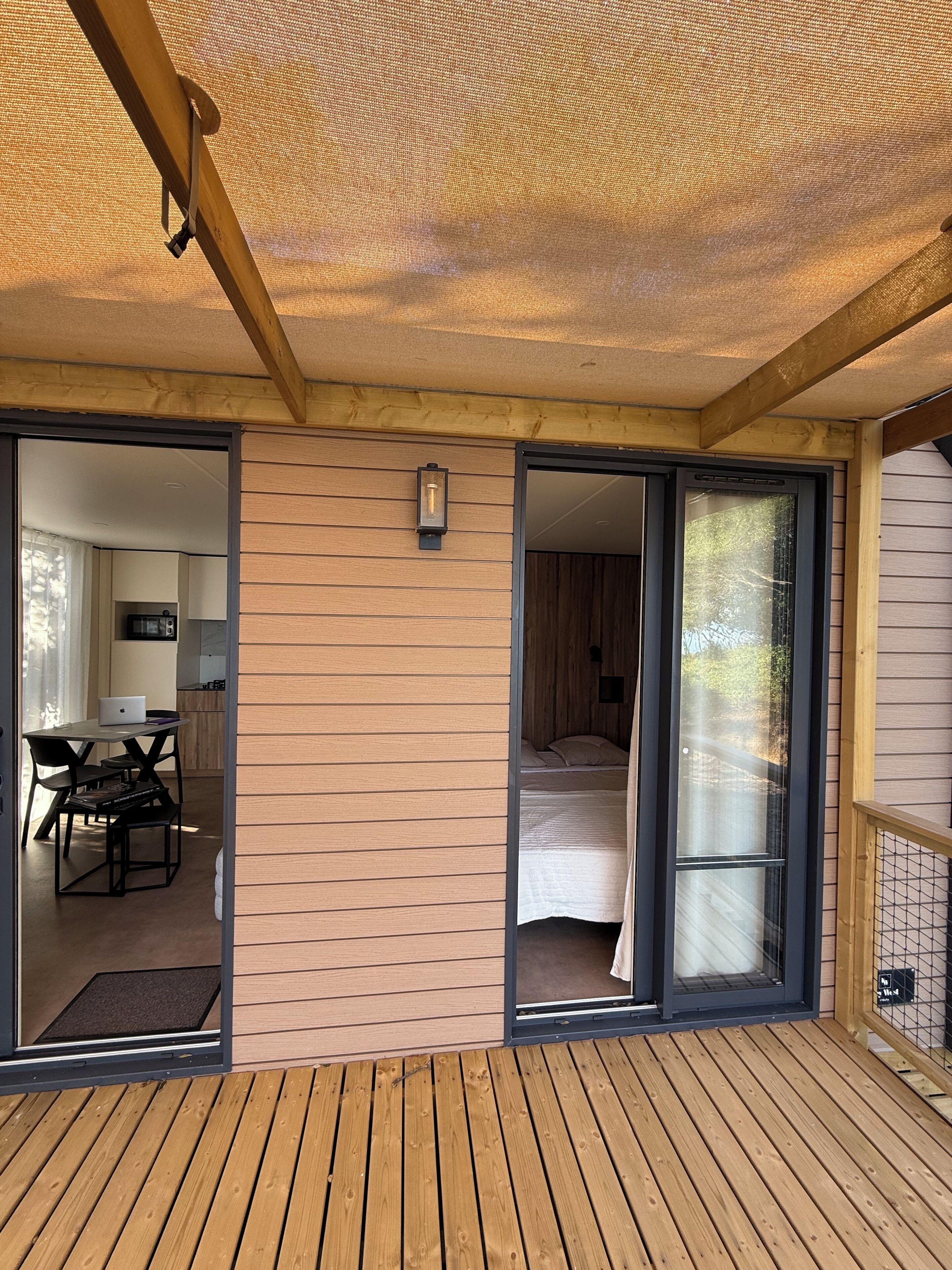 Una terraza de madera con un ventanal corredero que da a un dormitorio con cama y ventana.