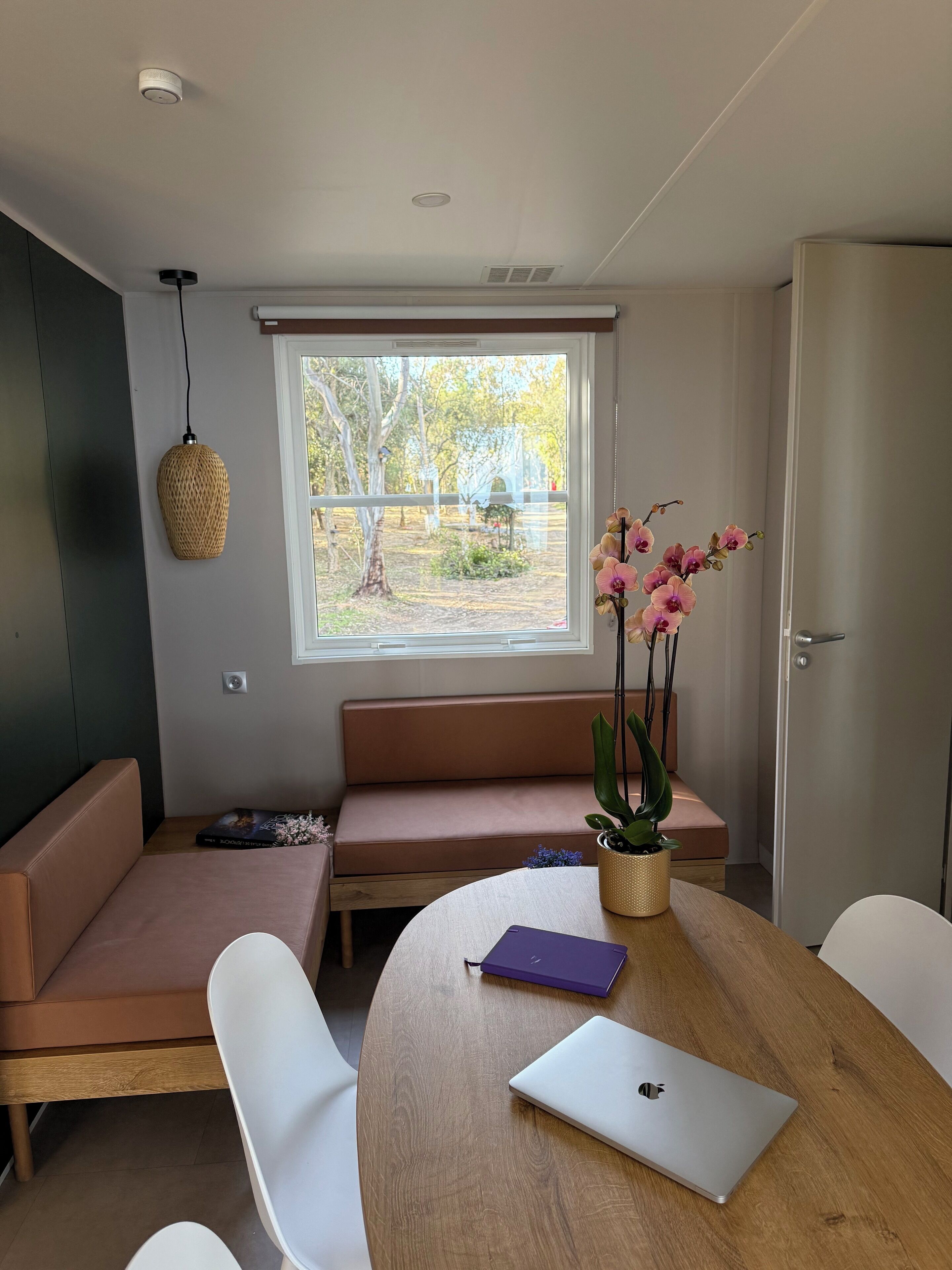 Ein modernes Zimmer mit einem Holztisch, einem Laptop, einer Couch und einem Fenster mit Blick auf Bäume und einen Zaun.